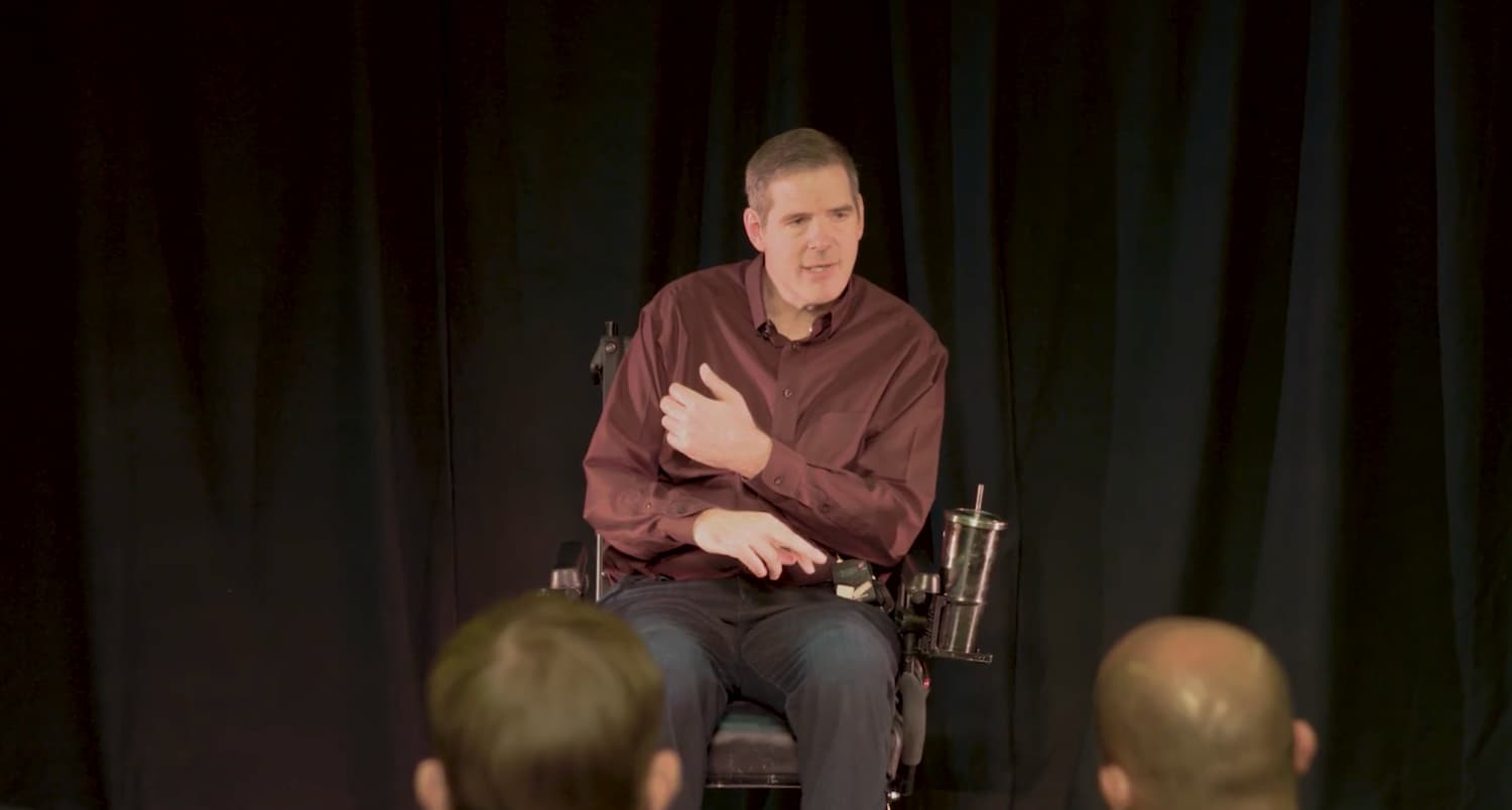 Scott Allen Frost speaking from his wheelchair to an audience, on a dark stage.